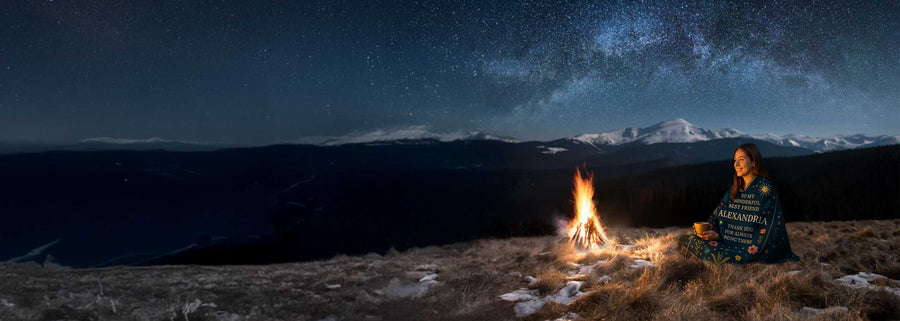 Girl wrapped in personalized best friend blanket sitting by campfire under starry night sky in the mountains