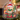 Personalized ugly Christmas sweater photo ornament — a real face above a garish knitted Christmas jumper and pom-pom hat in a cosy living room, on a red ribbon